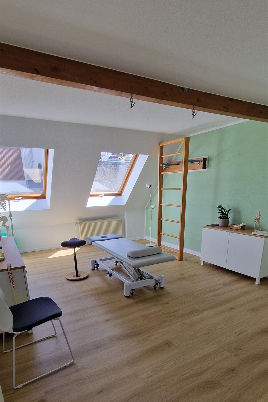 Heller Behandlungsraum mit zwei Dachfenstern, grauer Liege, Rollhocker und einer Sprossenwand vor salbeigrüner Wand.