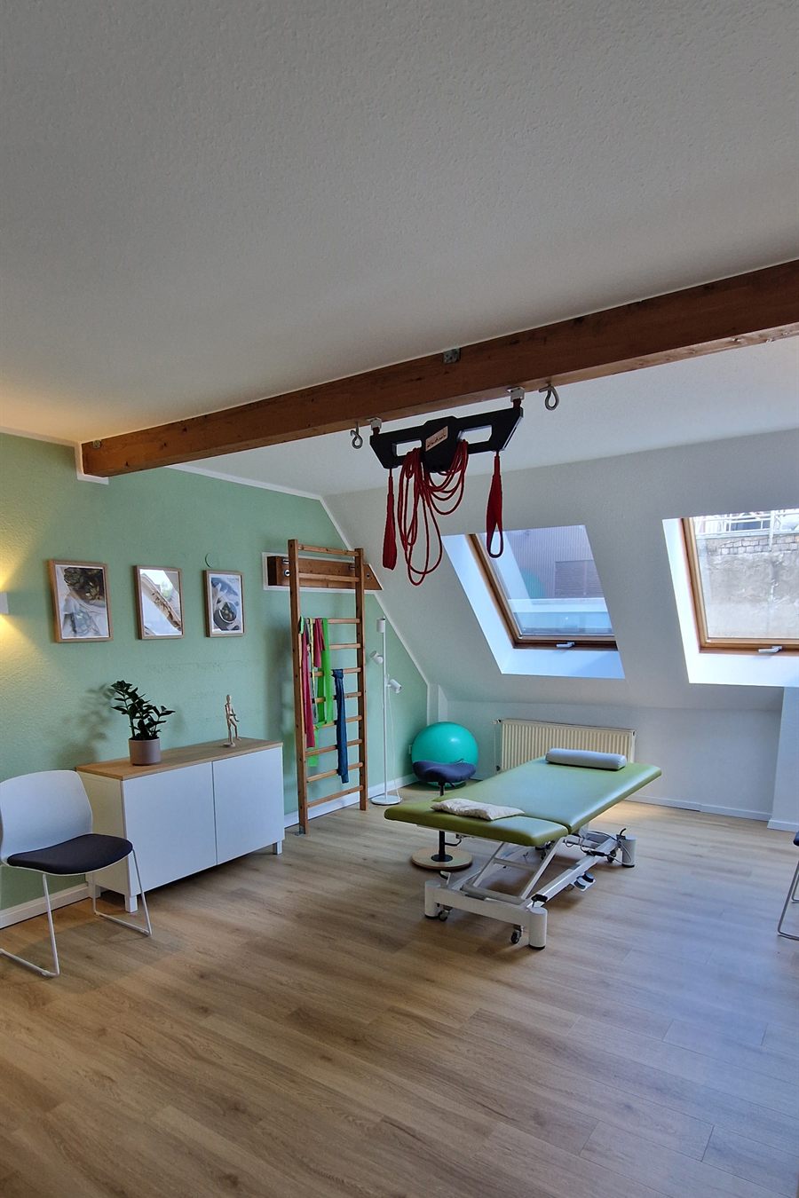 Therapieraum mit grüner Liege, Sprossenwand mit Therabändern, Gymnastikball und zwei Dachfenstern.