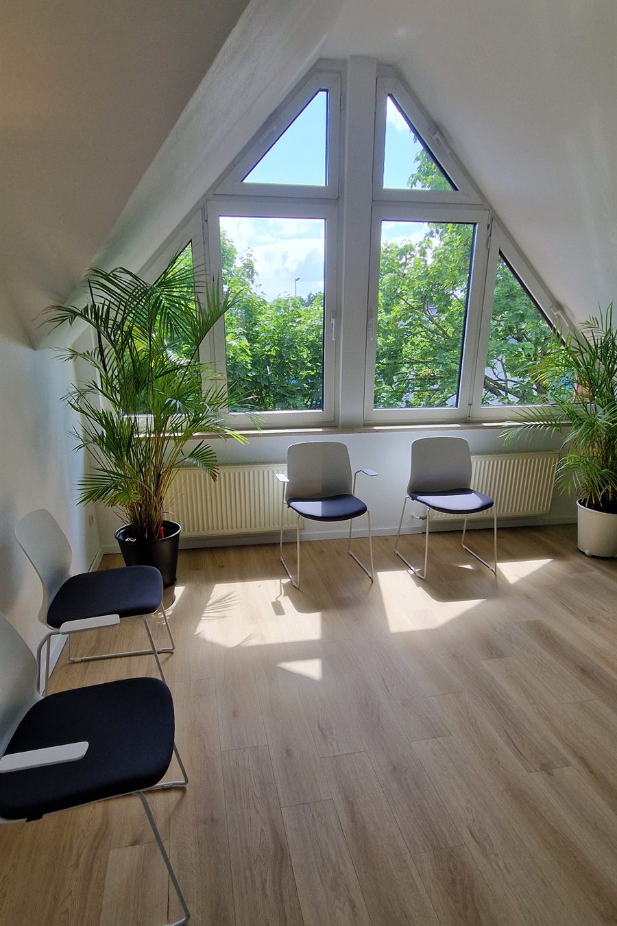 Wartebereich mit großem dreieckigem Giebelfenster, davor zwei Stühle, links eine Palme, Sonnenlicht auf dem Holzboden.